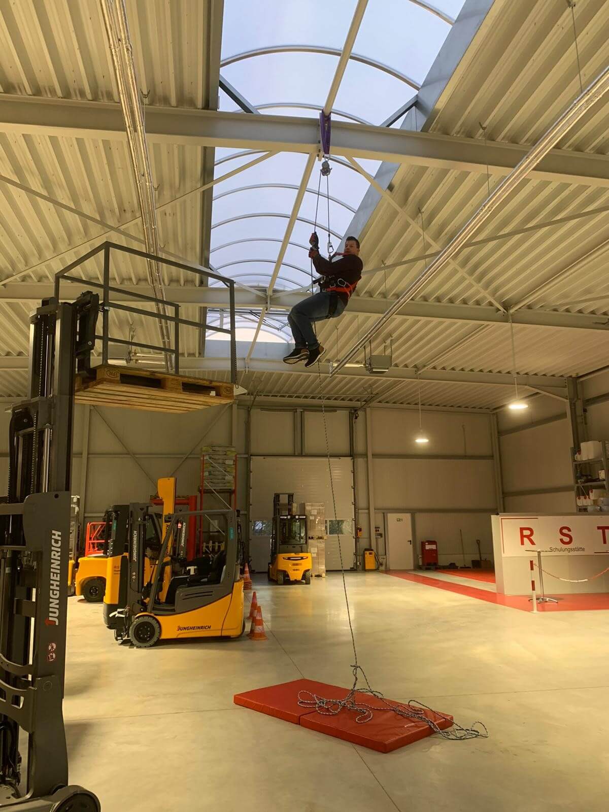 Große Lagerhalle mit Regalen voller Kartons, Gabelstaplern, Palettenstapel mit Leitkegel und rotem Hubwagen unter Dachfenster.