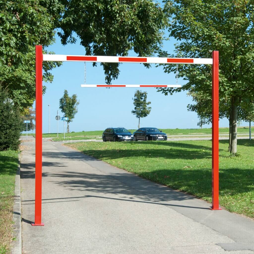 Roter und weißer Durchfahrtbegrenzer aus Metall auf gepflasterter Straße, zwei parkende Autos, Bäume im Hintergrund.