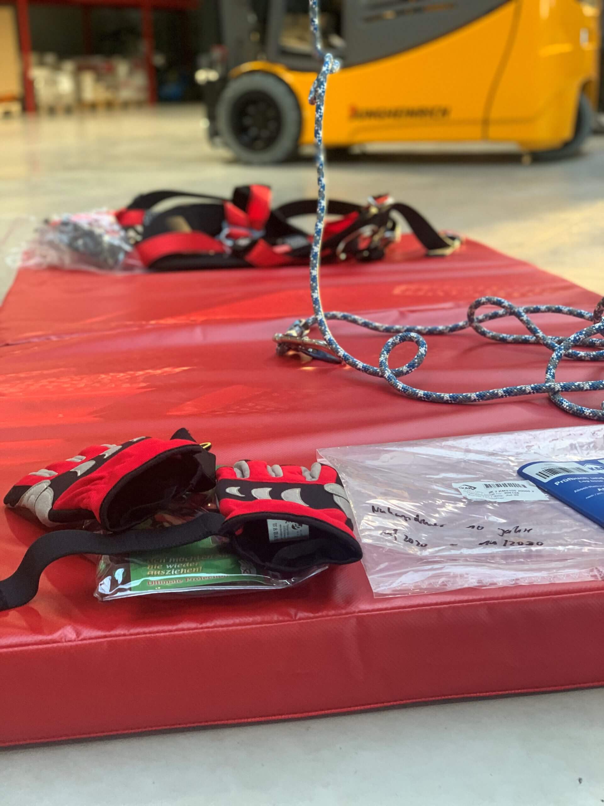 Ein Gabelstapler transportiert einen orangefarbenen Schutzkorb und Paletten vor einer Lagerhalle, die vier Personen in Warnwesten beobachten.