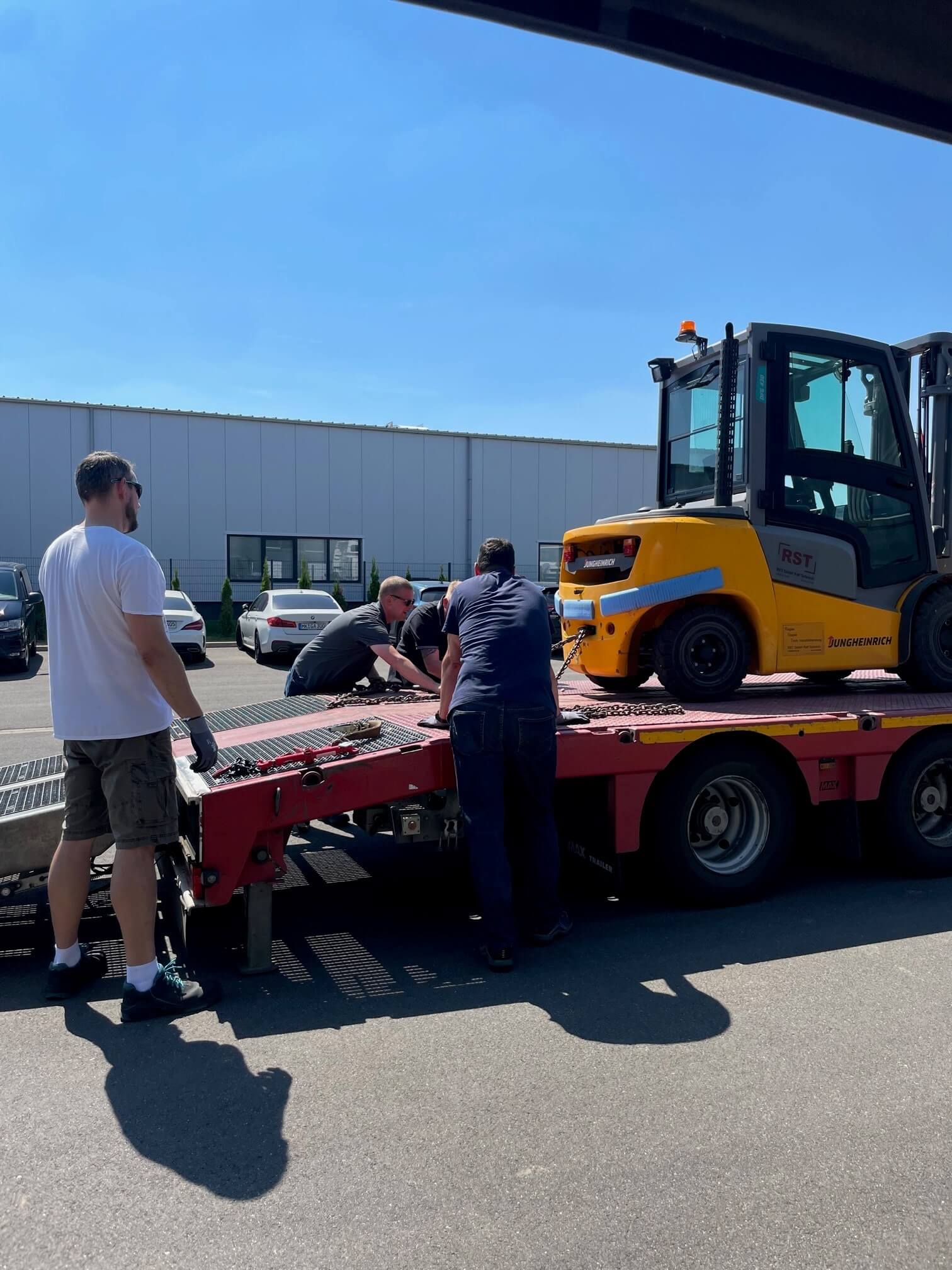 Zwei Personen mit Warnwesten stehen im Lagerhaus mit Holzpaletten, Regalen und Geräten unter einem Dach mit Oberlichtern.