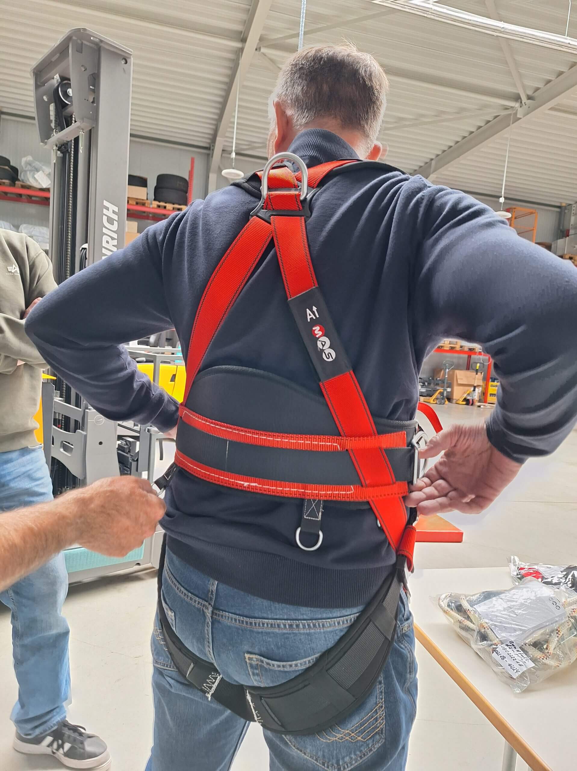 Back view of a man wearing a bright red safety harness being adjusted by another person in a workshop setting.