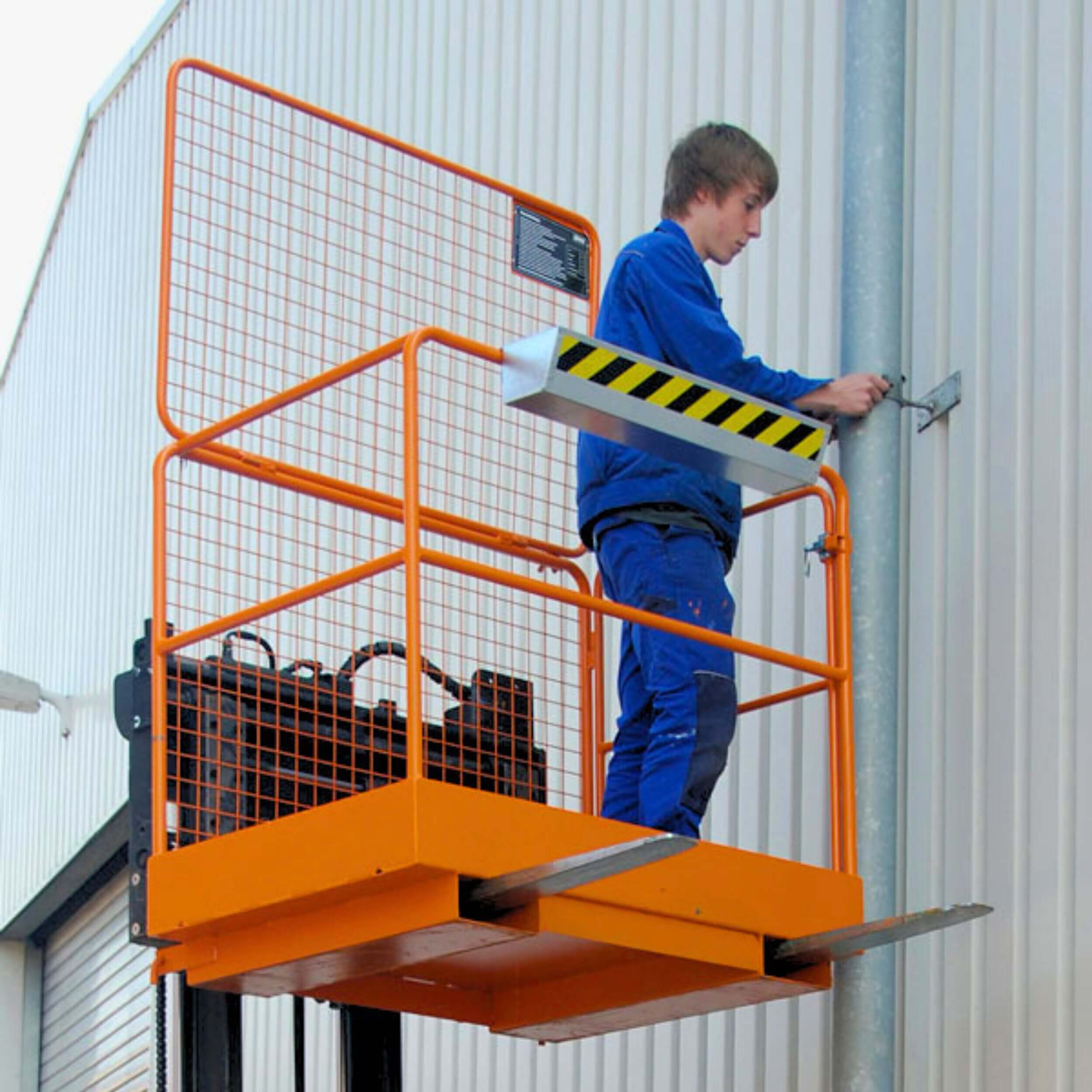Worker in blue overalls standing on an orange elevated work platform beside a metal building wall.