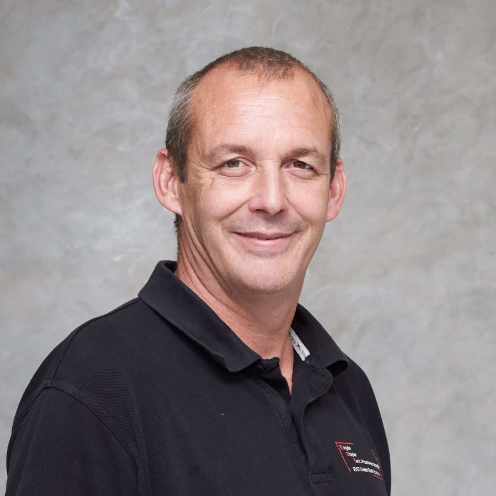Mann mit kurzem grauem Haar lächelt, trägt dunkles Hemd vor hellem Hintergrund; Porträt für Technische Industrieberatung.