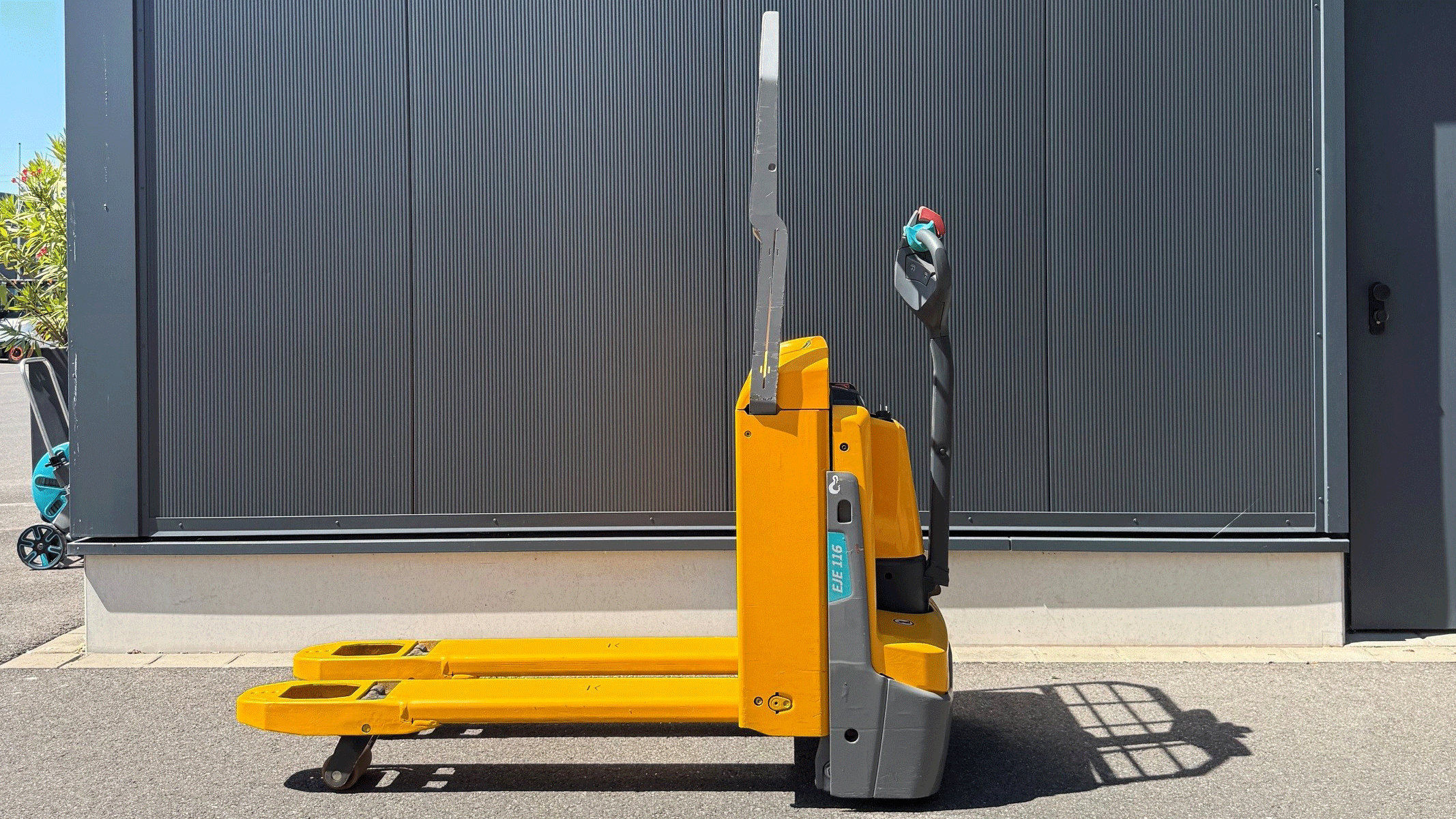 Yellow pallet jack with extended forks parked in front of a dark corrugated metal wall outside a building.