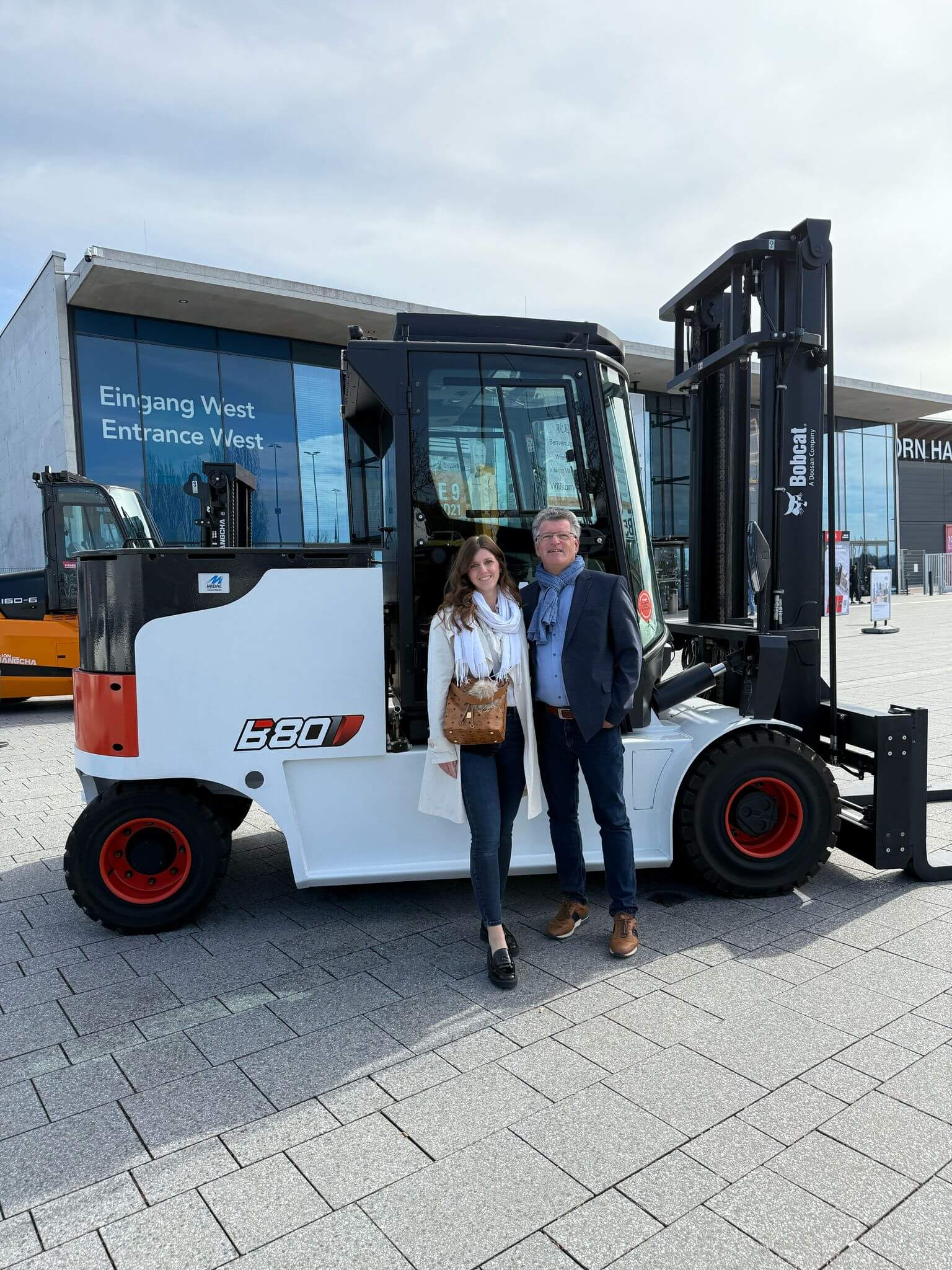 Ralf und Melina Schnirch vor der Messehalle Stuttgart (LogiMAT) und dem 8 Tonne Stapler von Bobcat, der auf der Messe ausgestellt wird.
