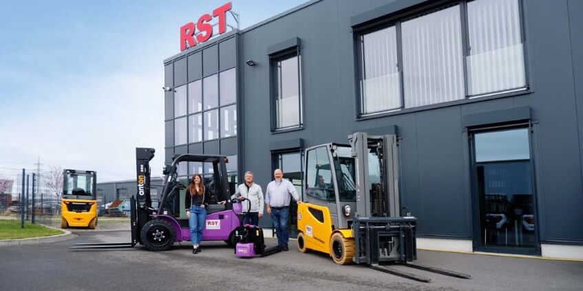 Group of four people standing beside a purple forklift and a yellow forklift outside a modern gray warehouse with an RST sign