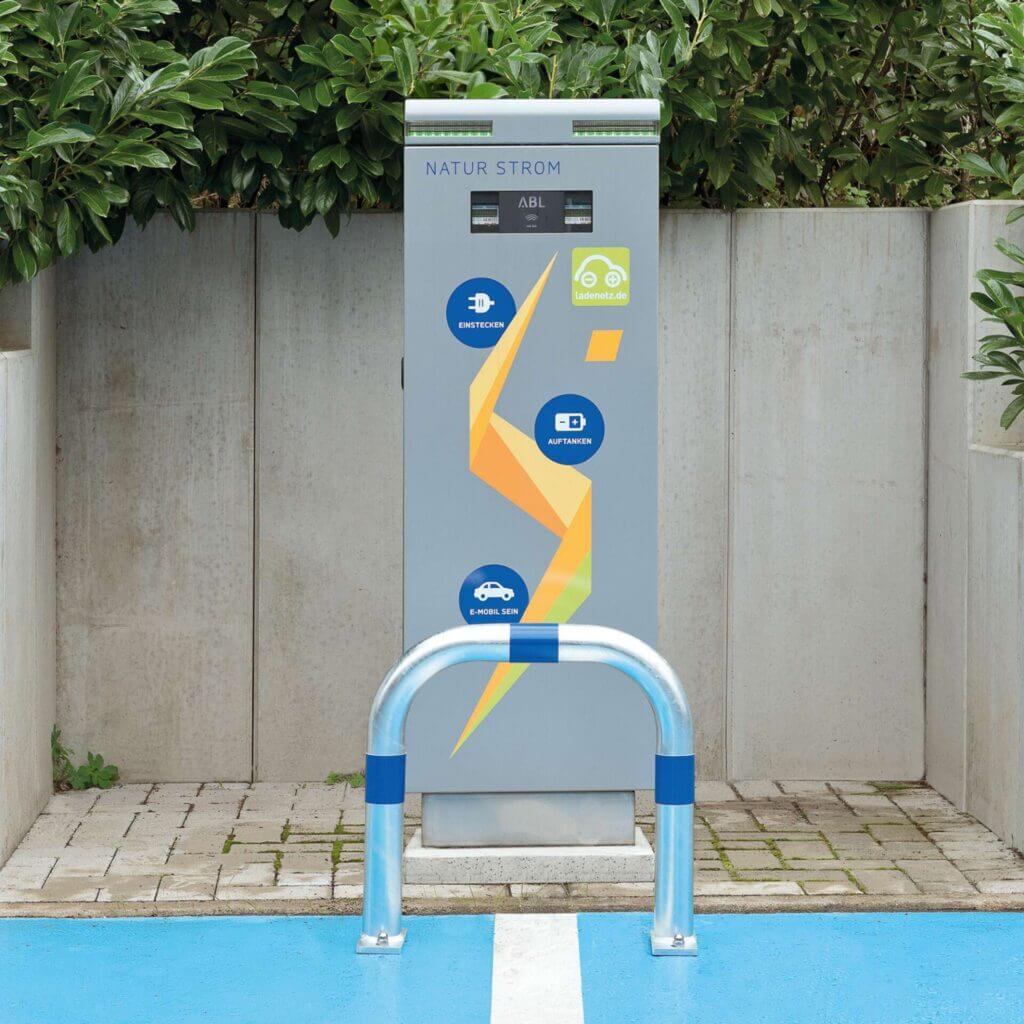 CITY-Schutzbügel GROß an E-Ladestation mit blauen und gelben Markierungen, umgeben von Grünfläche und Betonbarrieren.