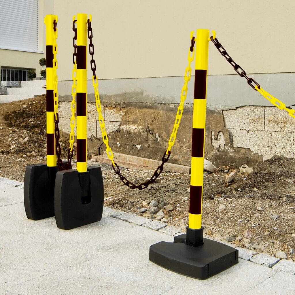 Drei gelb-schwarz gestreifte Ketten-Warnständer mit Kunststoffketten sperren eine Baustelle neben einem Gebäude ab.
