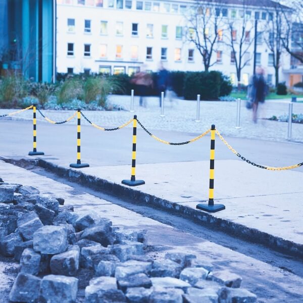 Das gelb-schwarze GUARDA Kettenständer-Set trennt einen ausgehobenen Bürgersteig mit losen Pflastersteinen vom angrenzenden Gehweg in einem städtischen Gebiet.