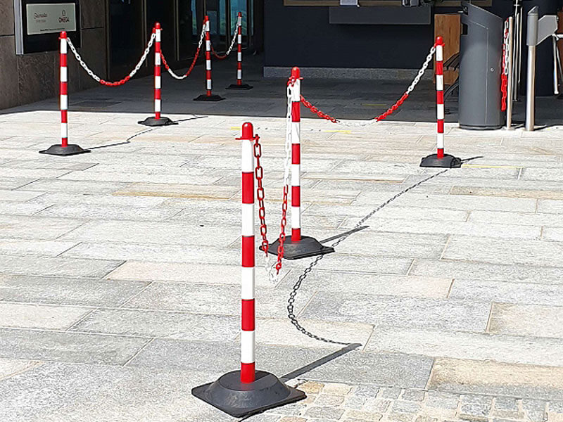 GUARDA Kettenständer-Set Ständer mit roten und weißen Streifen, die mit Ketten verbunden sind, bilden eine Barriere auf einem hellen Steinpflaster in der Nähe eines Gebäudeeingangs.