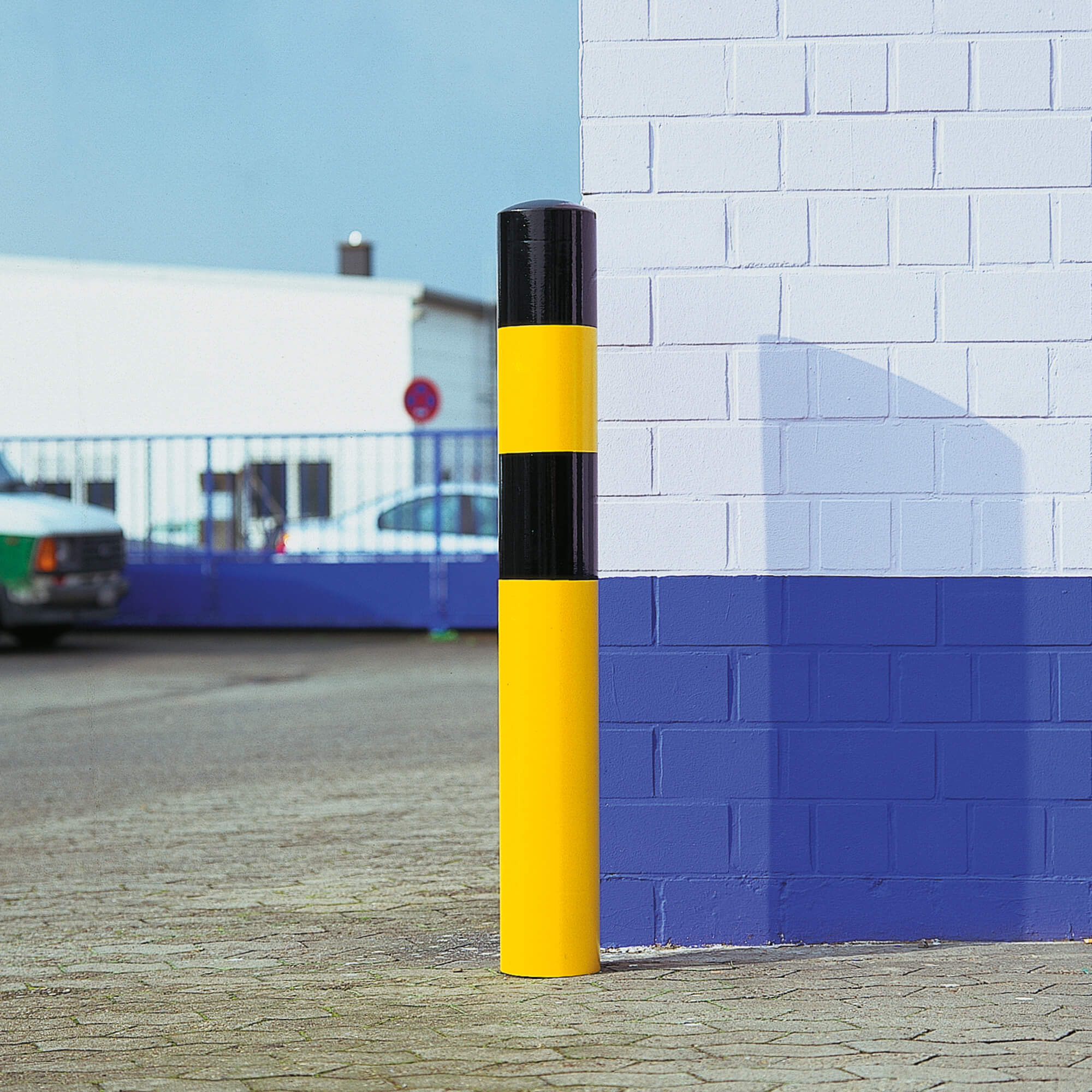 Yellow and black safety bollard in front of a blue wall in an industrial area.