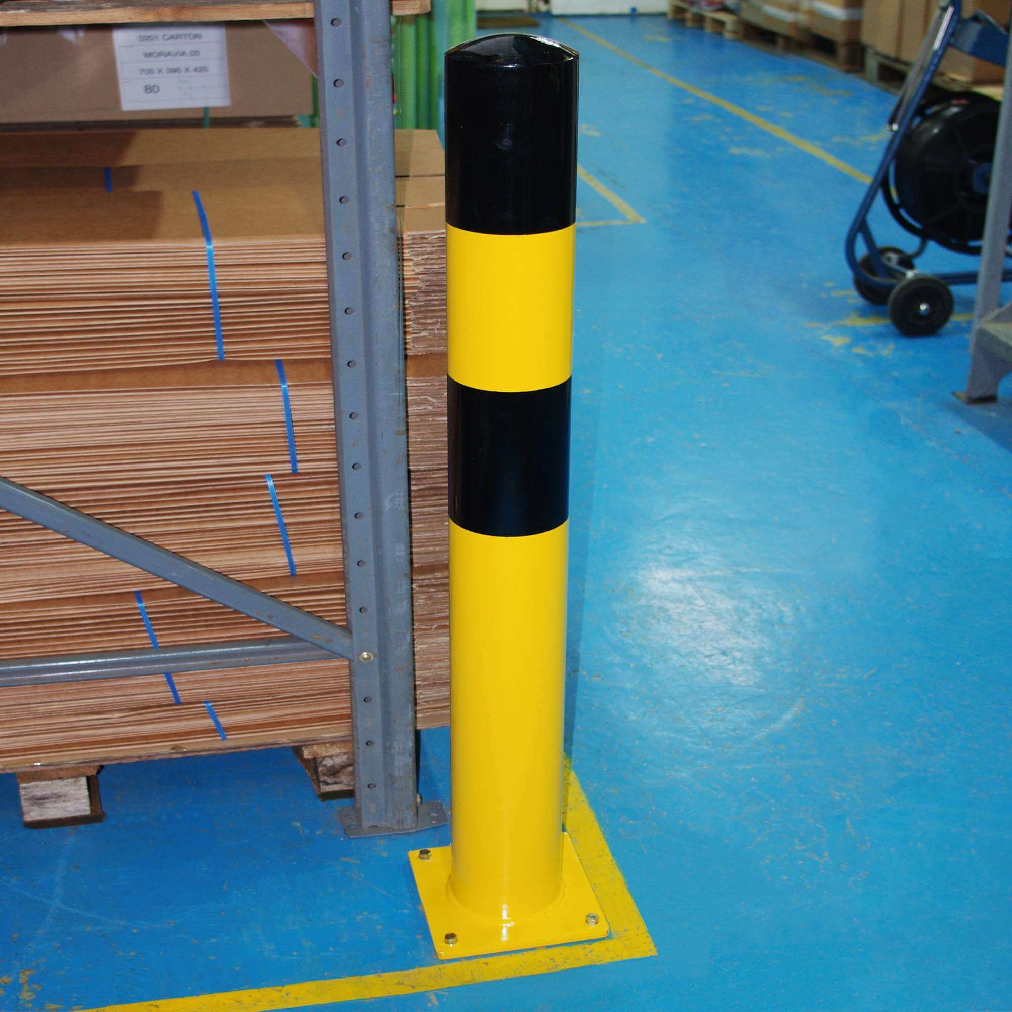 Yellow and black safety bollard protecting a rack in a warehouse, with stacked plywood on the left and a blue floor with yellow lines.