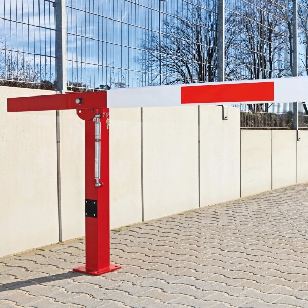 Eine COMPACT Handschranke in Rot und Weiß ist auf einer gepflasterten Fläche neben einem hohen Drahtzaun und einer Betonmauer installiert.