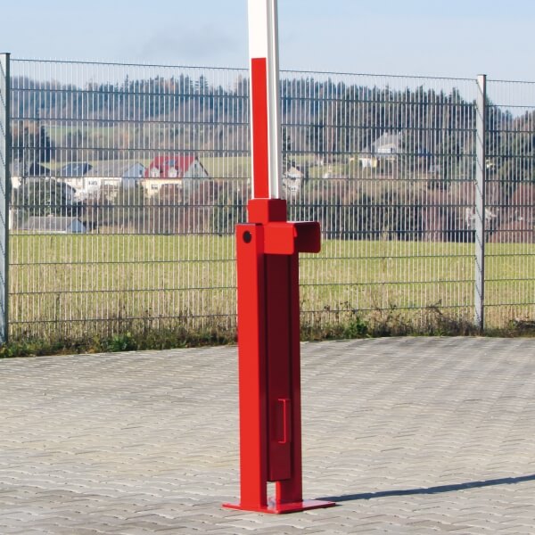 Eine COMPACT Systemschranke mit Gegengewicht in Rot und Weiß steht auf einer gepflasterten Fläche, mit einem Metallzaun und einer Wiese im Hintergrund.