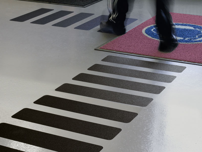 Person läuft auf Boden mit schwarzen Antirutschstreifen, die zu roter Matte mit blauem Kreis führen; rutschfester Bodenbelag.
