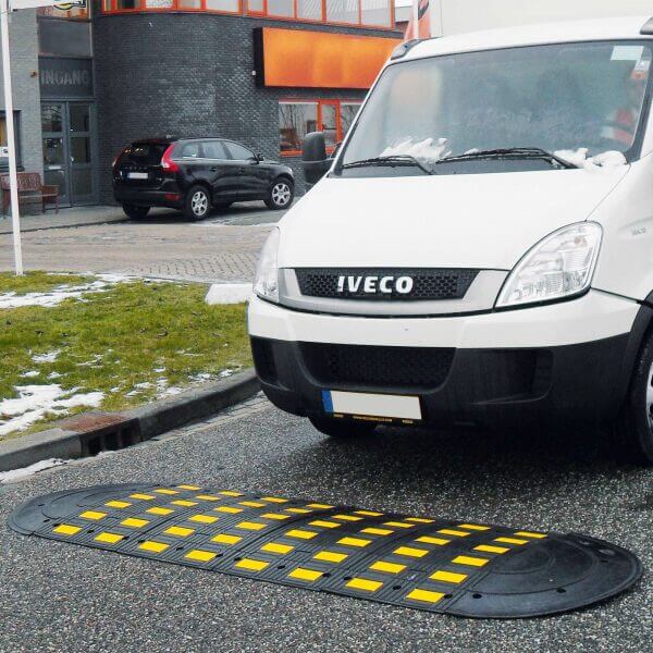 Weißer Iveco Lieferwagen an Straße, neben großem gelb-schwarzem SafeRide Bremsschweller, Wiese und Gebäude im Hintergrund.