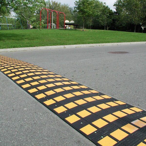 Gelb-schwarze SafeRide Bodenschwelle auf Asphaltstraße, daneben Park mit Gras, Bäumen und Spielplatzgerät im Hintergrund.