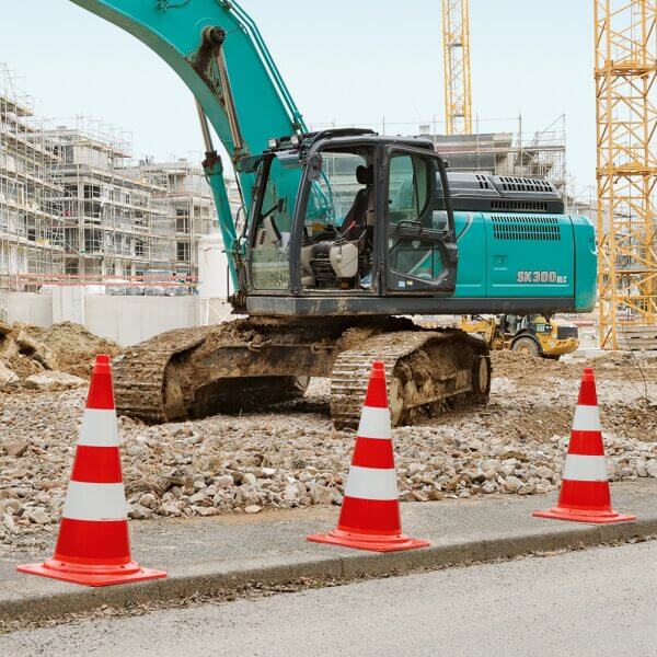 Türkiser Bagger auf Schotter am Bauplatz, davor vier MORION-Leitkegel-EU, im Hintergrund Gerüst und Baukräne.