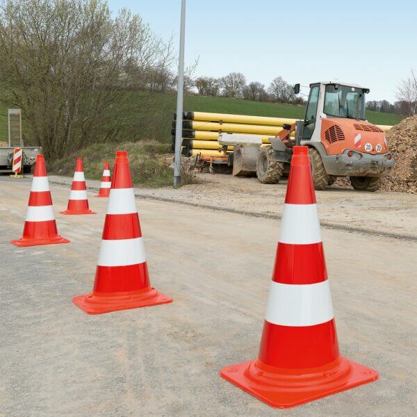Vier MORION-Leitkegel EU orange-weiß stehen an Straße bei Bauarbeiten, gelbe Rohre, Erdanhäufung und Bäume im Hintergrund.