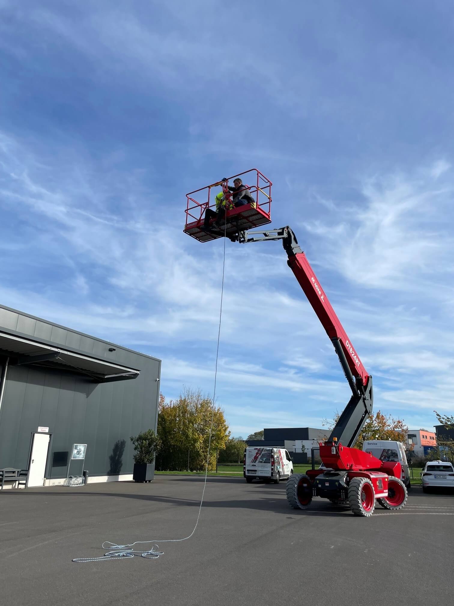 Ein Gabelstapler transportiert Holzpaletten vor einem Lagerhaus, mehrere Personen mit Warnwesten beobachten bei klarem Wetter.