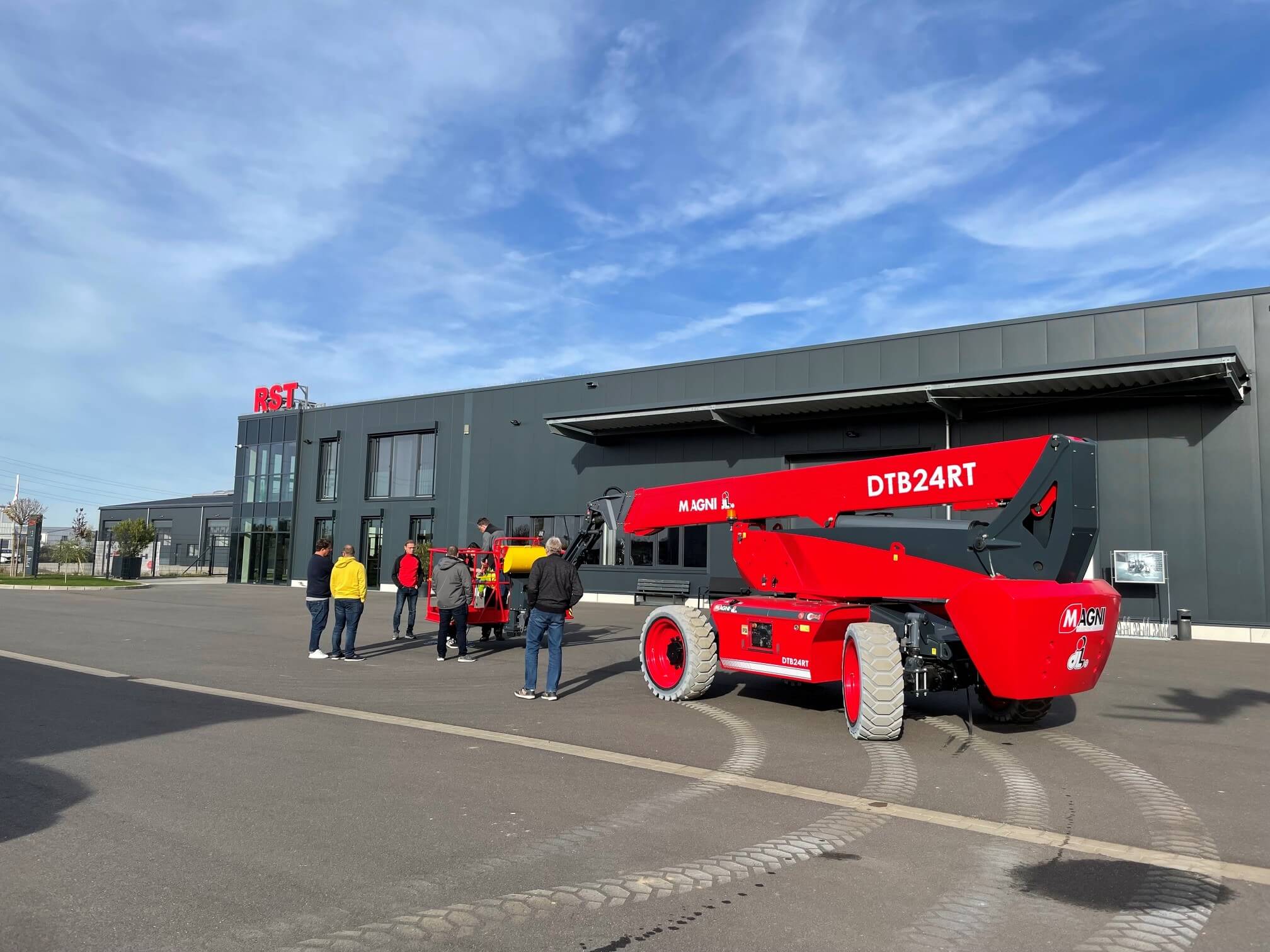 Ein Gabelstapler transportiert einen orangefarbenen Schutzkorb und Paletten vor einer Lagerhalle, die vier Personen in Warnwesten beobachten.