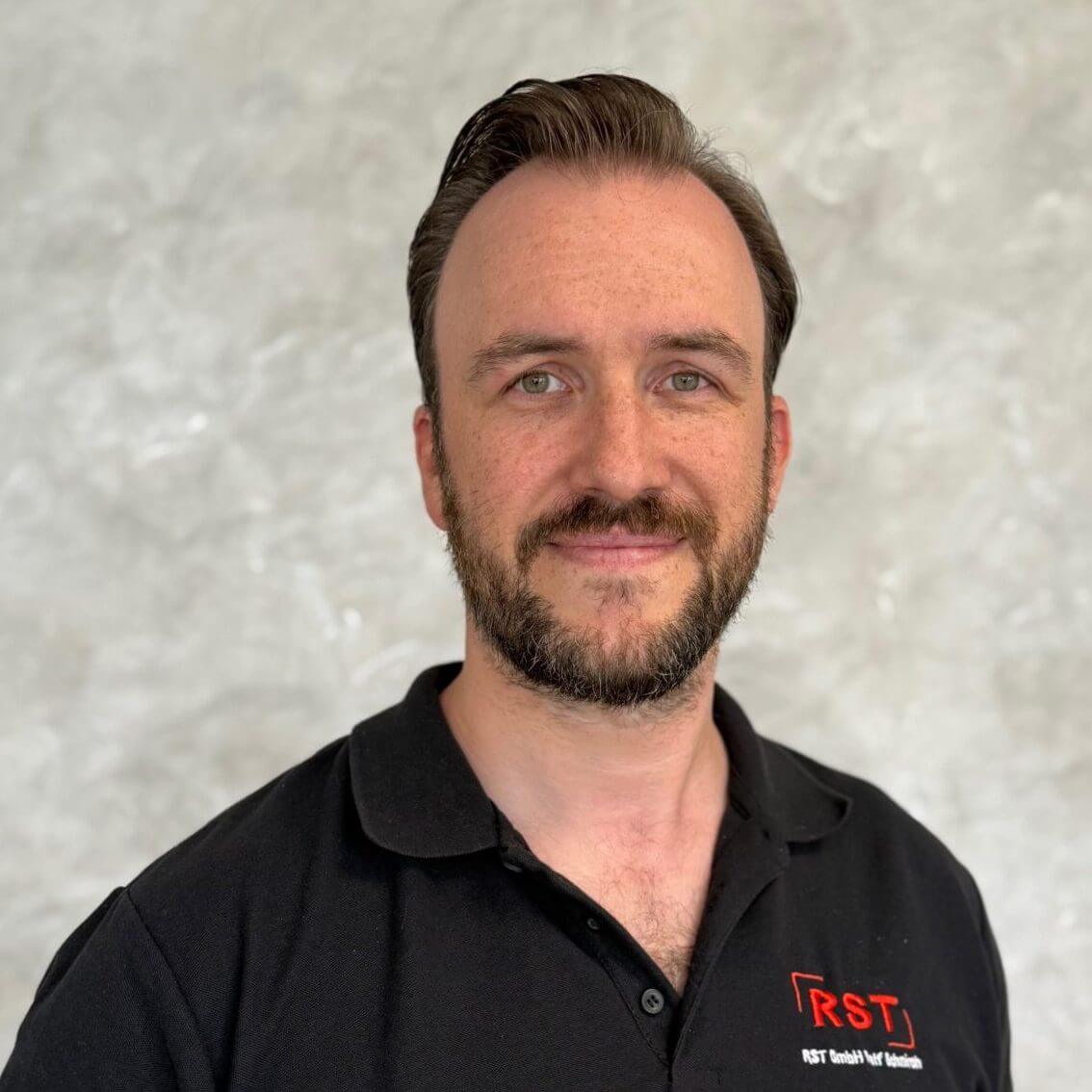 Mann mit kurzem grauem Haar lächelt, trägt dunkles Hemd vor hellem Hintergrund; Porträt für Technische Industrieberatung.