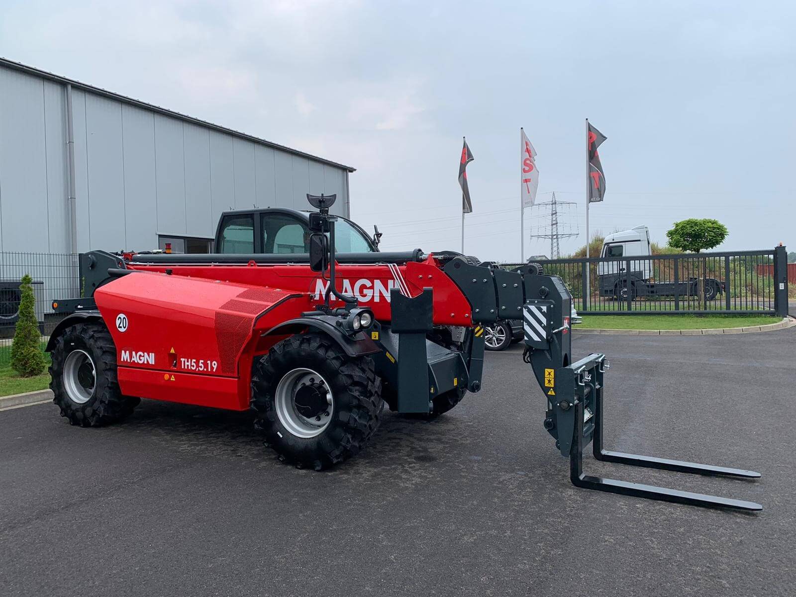 Red Magni telehandler with pallet forks attached, parked on a paved lot beside an industrial building.