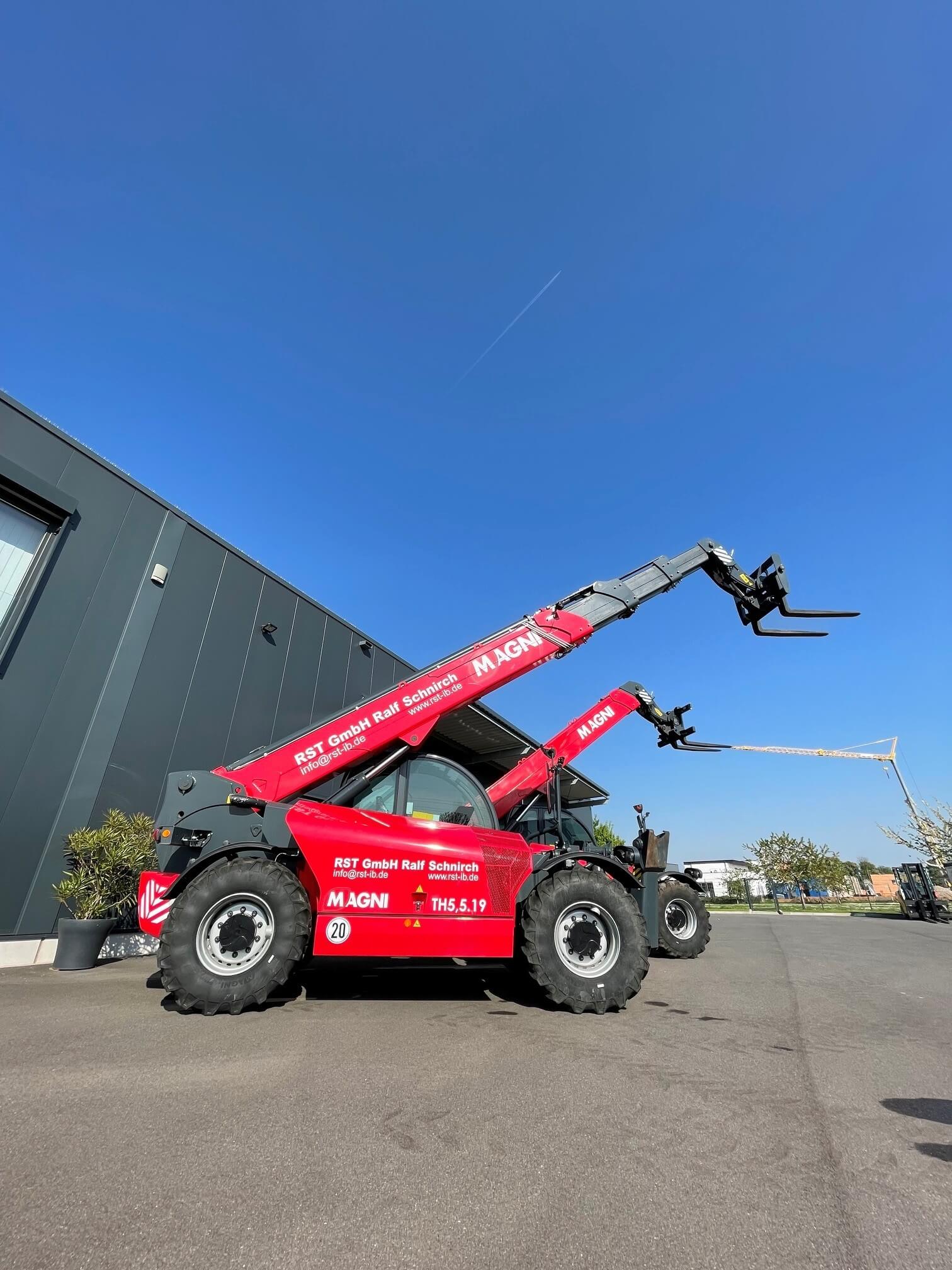 Red Magni mobile crane with its lifting arm extended, parked beside a dark gray industrial building under a clear blue sky. Note: brand 'MAGNI' visible on the crane arm.