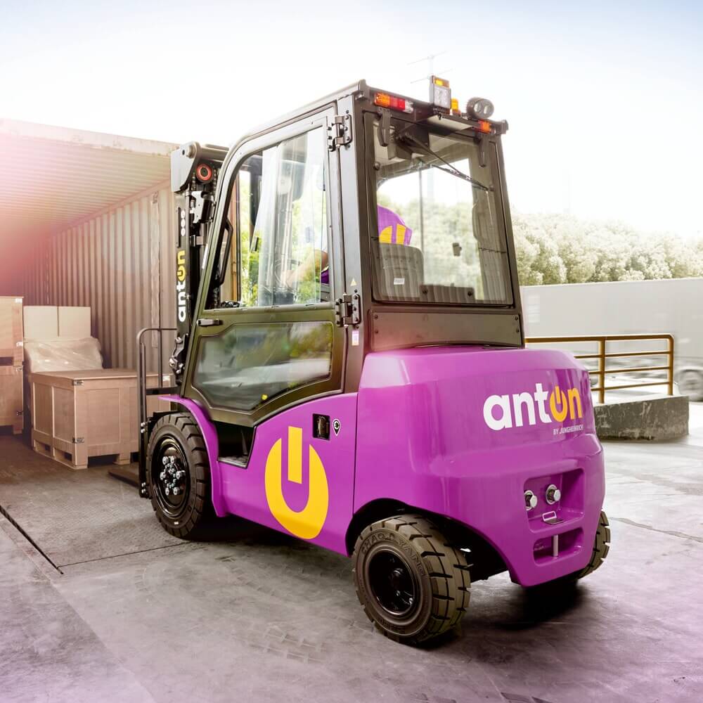 Ein AntOn von Jungheinrich Elektro-Stapler CBH 2.5 mit einer Hubhöhe von 4.800 mm und Wetterschutzkabine Com 3 entlädt an einer Laderampe Holzkisten aus einem Versandcontainer.