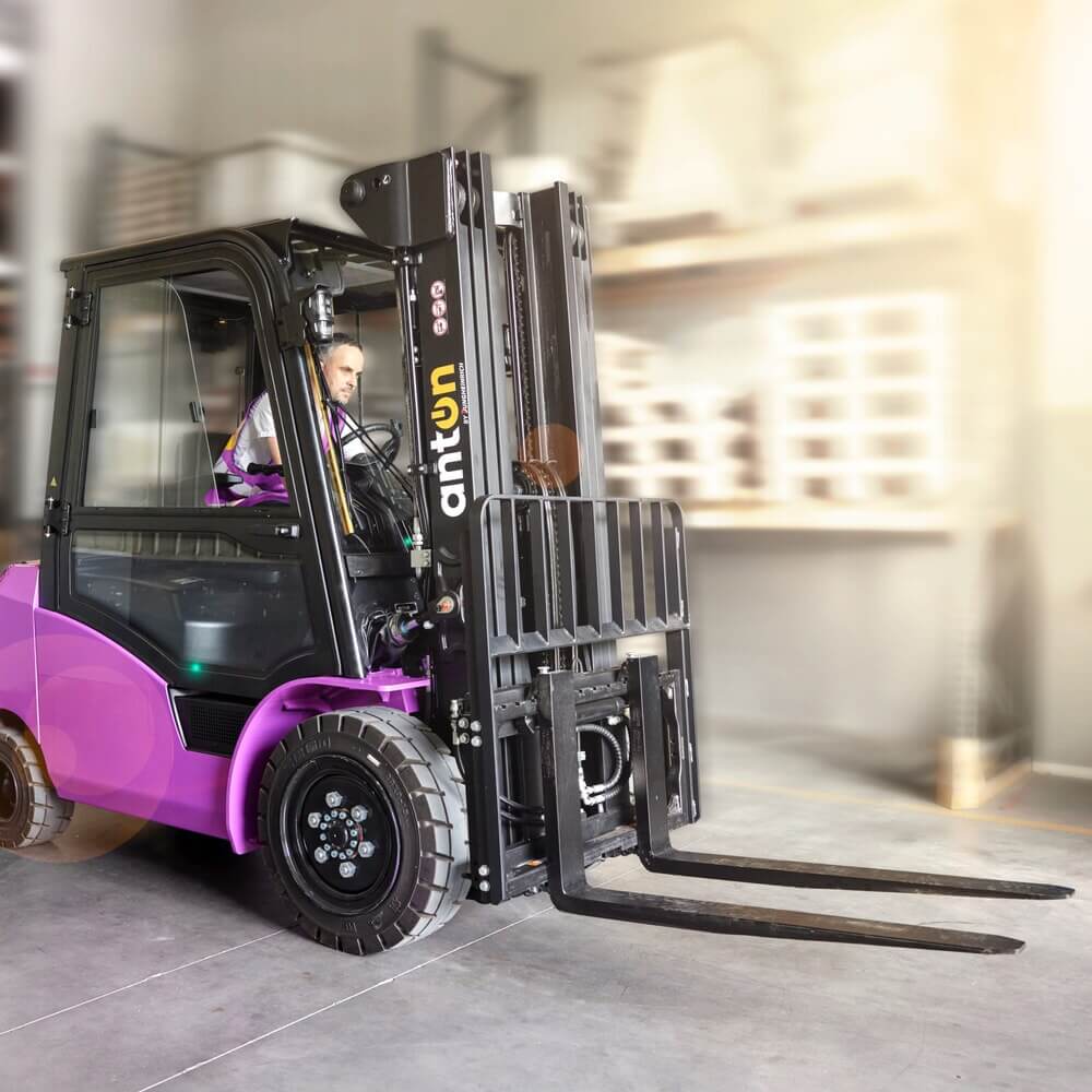 Eine Person bedient einen violetten AntOn von Jungheinrich Elektro-Stapler CBH 2.5 mit Hubhöhe 4.800 mm und Wetterschutzkabine Com 3 in einem Lagerhaus, umgeben von Regalen und verschwommener Beleuchtung im Hintergrund.