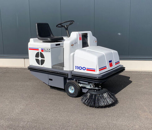 Aufsitz-Kehrmaschine mit Frontbürste steht auf Pflaster vor grauer Wand; Industriereiniger, Bodenreinigung, Kehrfahrzeug.