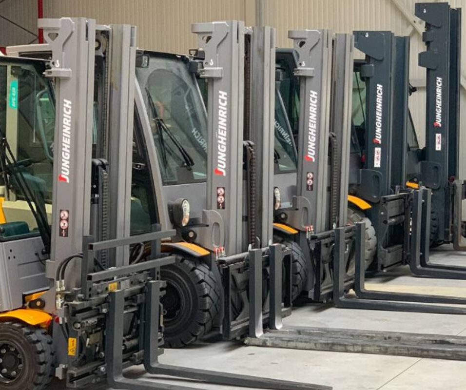 Vier Jungheinrich Gabelstapler stehen nebeneinander in einer Lagerhalle, Gabeln angehoben und parallel ausgerichtet.