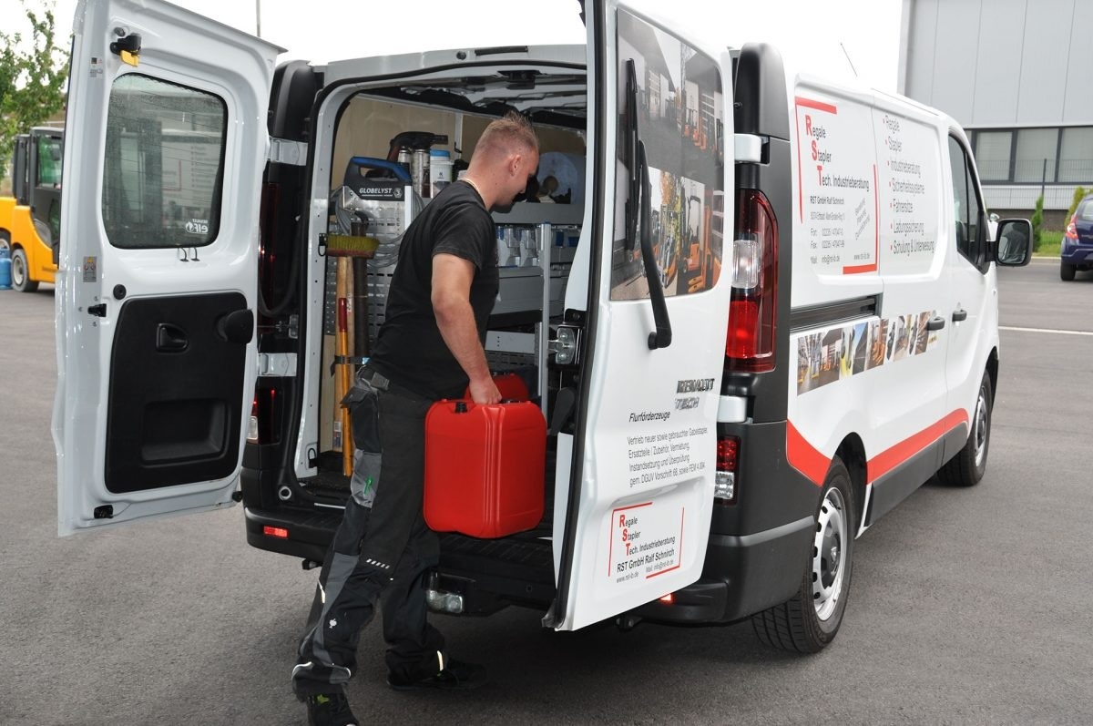Ein Mann lädt einen roten Benzinkanister in einen weißen Werkstattwagen mit Werkzeugen auf einen gepflasterten Parkplatz.